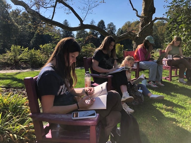 A group of students sits on chairs and on the ground in a garden while writing.