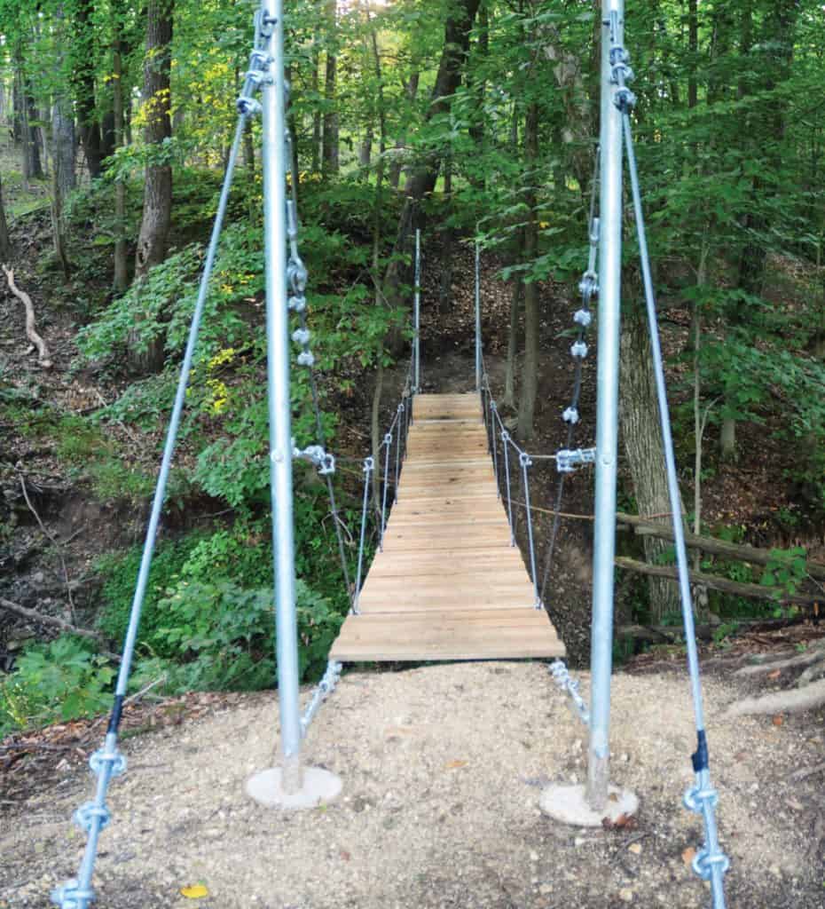 A wooden and metal suspension bridge crosses a creek in the shady forest at ChesLen Preserve.