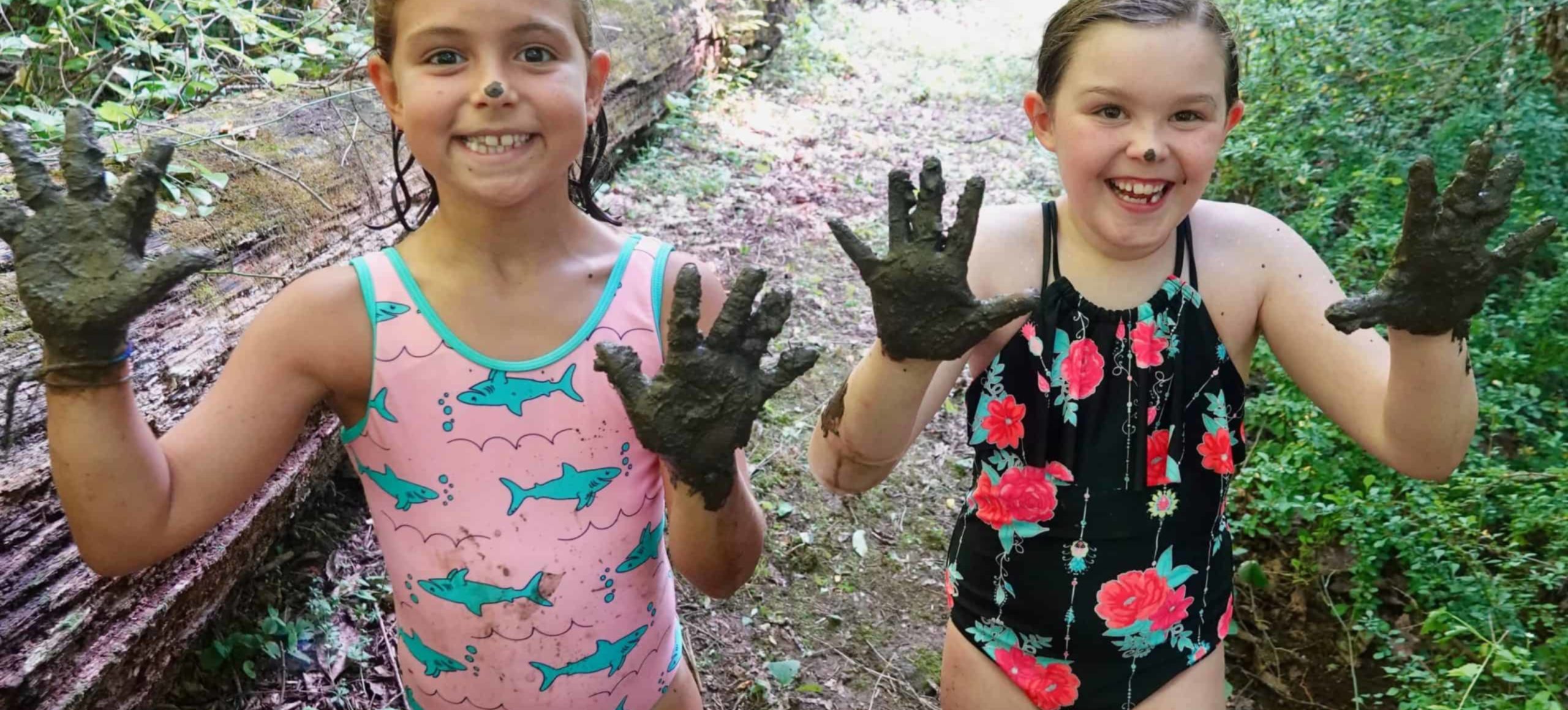 Two girls wearing bathing suits face the camera. They're holding up muddy hands and have dollops of mud on their noses. Big smiles.