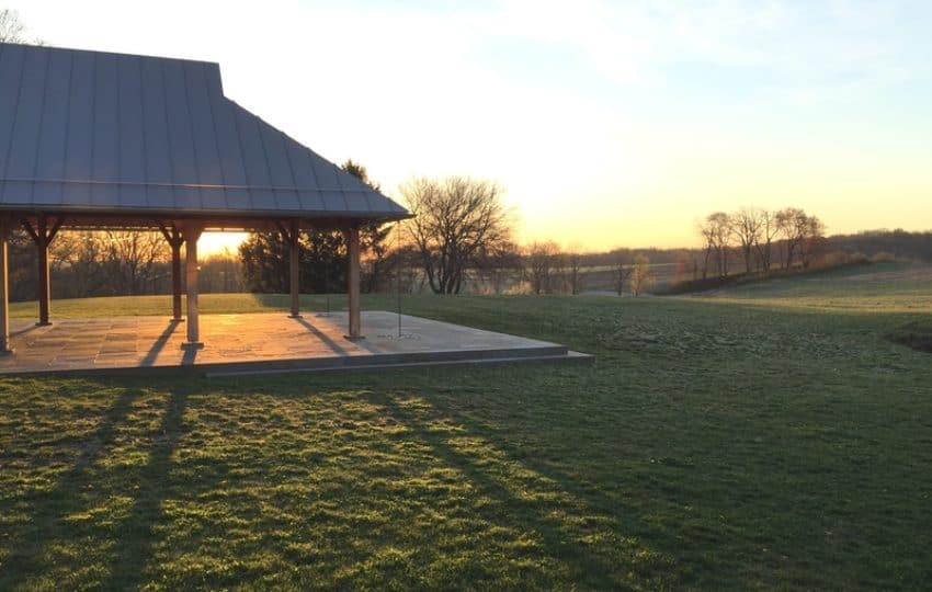 a pavilion sits in a large green field with its shadow cast on the grass as the sunrise shines behind it.