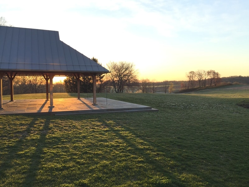 a pavilion sits in a large green field with its shadow cast on the grass as the sunrise shines behind it.