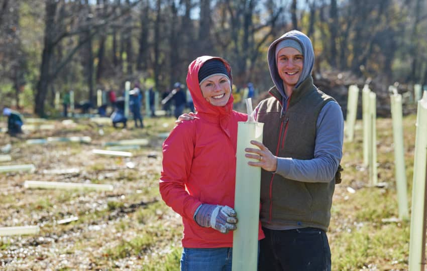 A woman with a red hooded puffy jacket and a man in a hoodie and vest smile as they place a tree tube in a field.
