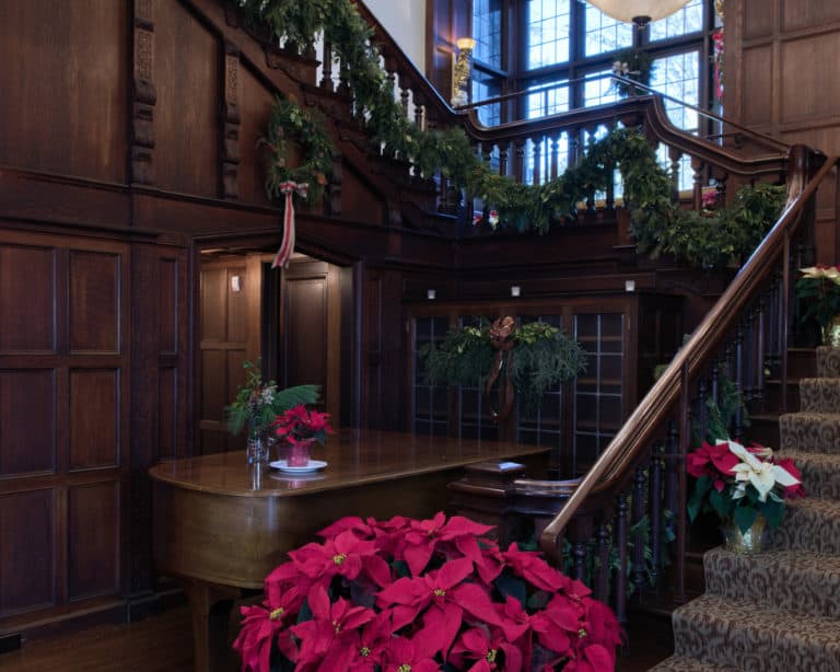 A bright red poinsettia sits at the bottom of a staircase, a lit chandelier above the stairs midway and evergreen boughs wrapped around the staircase.
