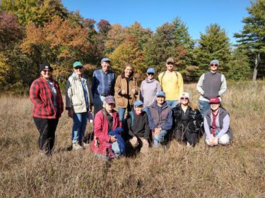 A smiling group of three Natural Lands staff and nine volunteers at a seed collection day, the field is brown grasses and behind them a long row of trees with a variety of fall colors - red, orange, yellow, green