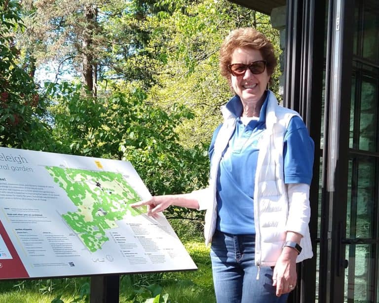 Smiling woman in sunglasses pointing to a sign with a map of Stoneleigh garden