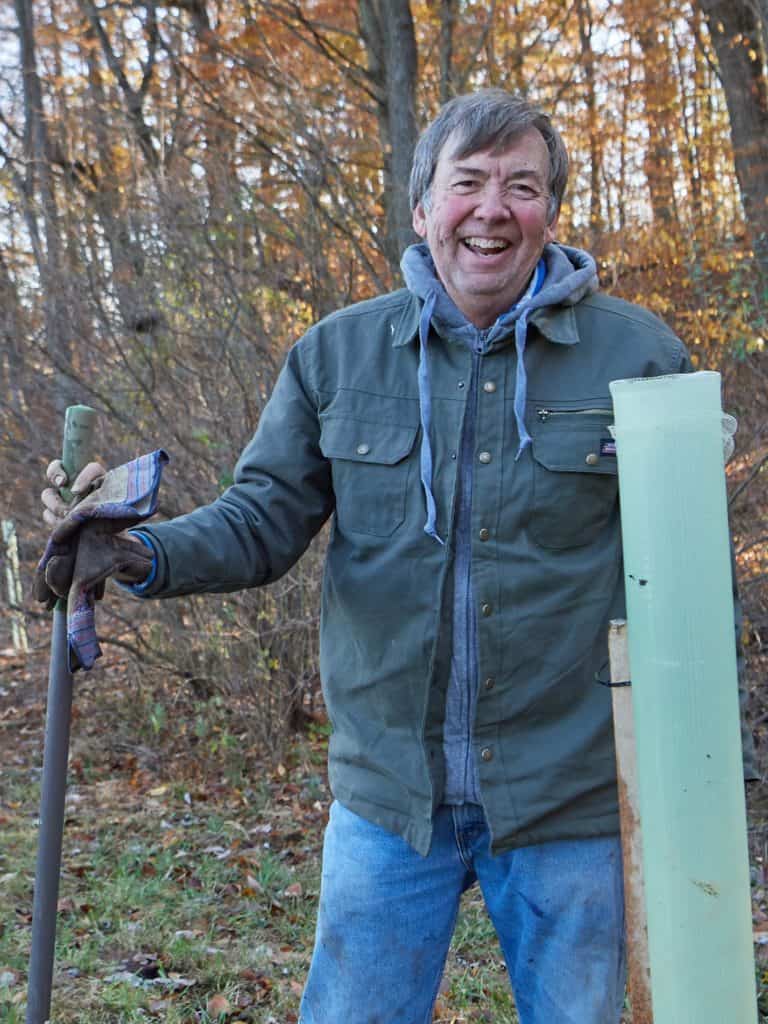 A man volunteer standing next to a tree tube near a forest, smiling