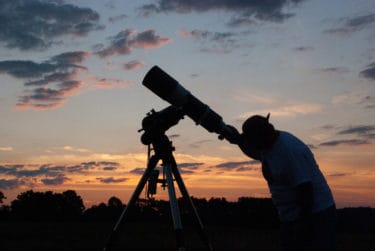 The silhouette of a person looking up through a telescope with a vibrant orange and blue sunset in the background.