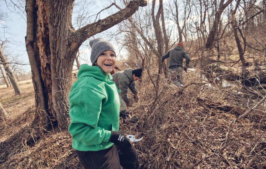 A person in a green hoodie and gray knit cap smiles at the camera while two other volunteers are in the background.