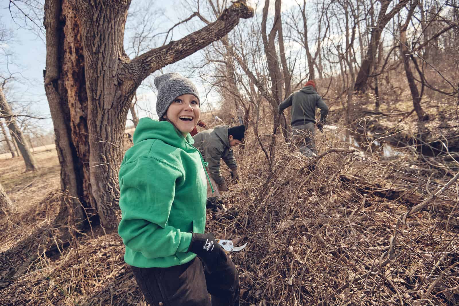 A person in a green hoodie and gray knit cap smiles at the camera while two other volunteers are in the background.