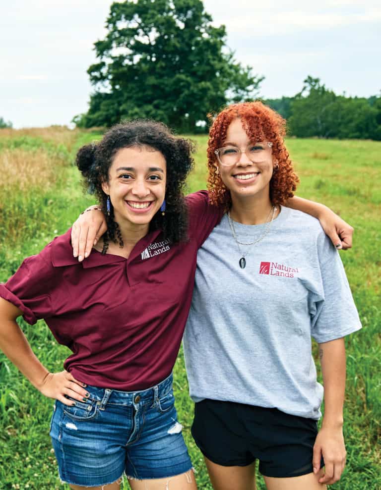 Two young women stand with arms around each others' shoulders in a green meadow