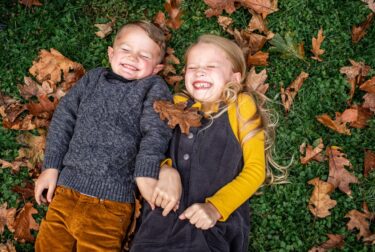 A girl and a boy in fall clothing smile with their eyes closed while lying on green grass and brown autumn leaves.