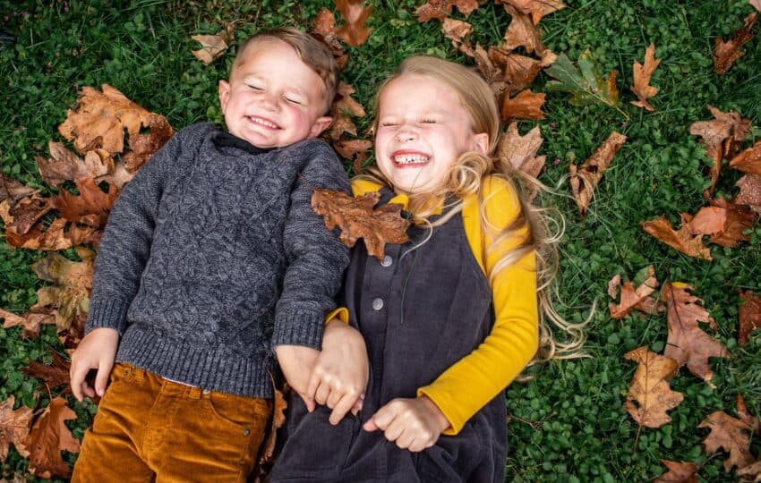 A girl and a boy in fall clothing smile with their eyes closed while lying on green grass and brown autumn leaves.