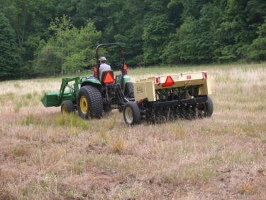 A man drives a green tractor with a no-till drill planting seeds in a brown meadow. 