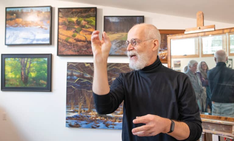 A man in a black turtleneck gestures with his hands while talking. He is in front of a wall of paintings.