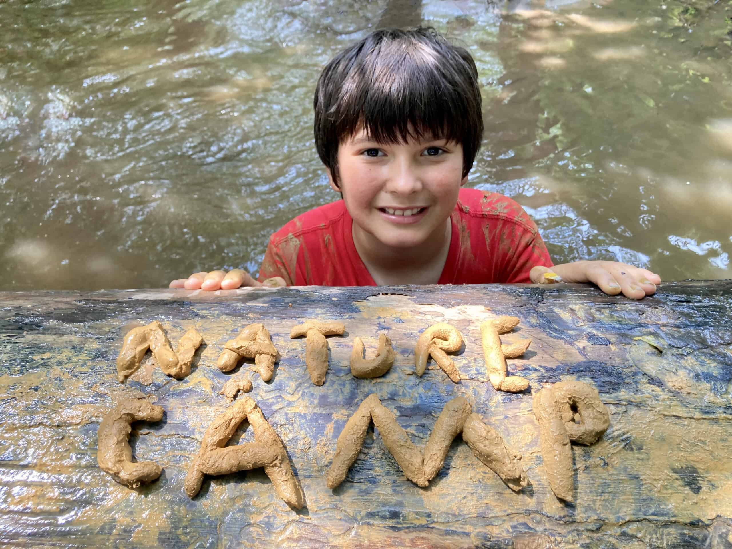 Camper posing with the words Nature Camp spelled out in pieces of rolled clay on a log in a creek