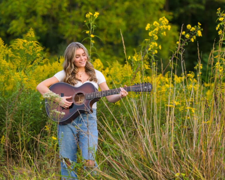 A woman wearing jeans and a t-shirt stands in a meadow of flowers and high grass playing a guitar.