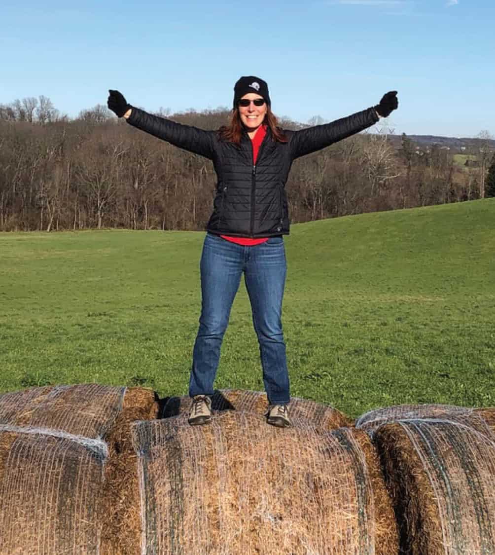 A woman in a black coat and hat standing on top of hay bales in a green field.