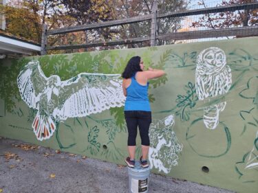 Sienna Proetto stand on a bucket to paint a mural at Binky Lee Preserve