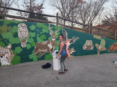 Sienna carries a baby in a carrier on her back while standing in front of her unfinished mural of plants and animals.