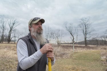 Man with hat, beard, an vest looking into the distance, standing in field with trees.