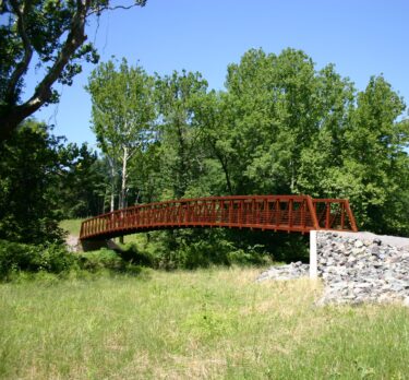 A steel bridge spans a creek with green grass and tall, leafy trees on both sides. 