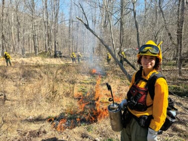 A woman in fire protective gear holds a torch in front of a controlled fire in a meadow.