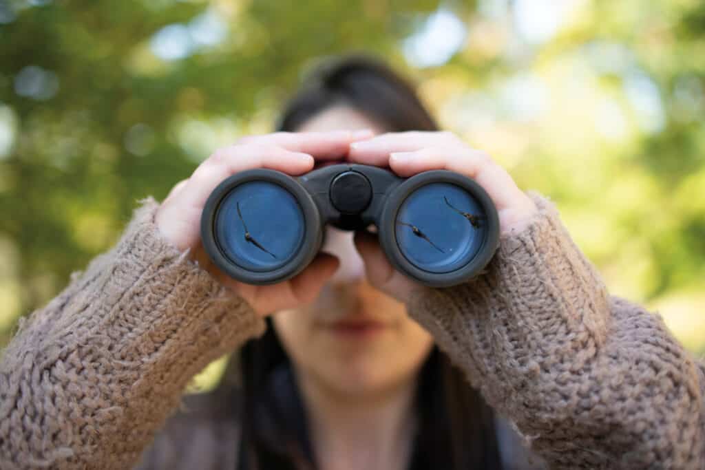 A woman in a tan sweater looks through binoculars outside.