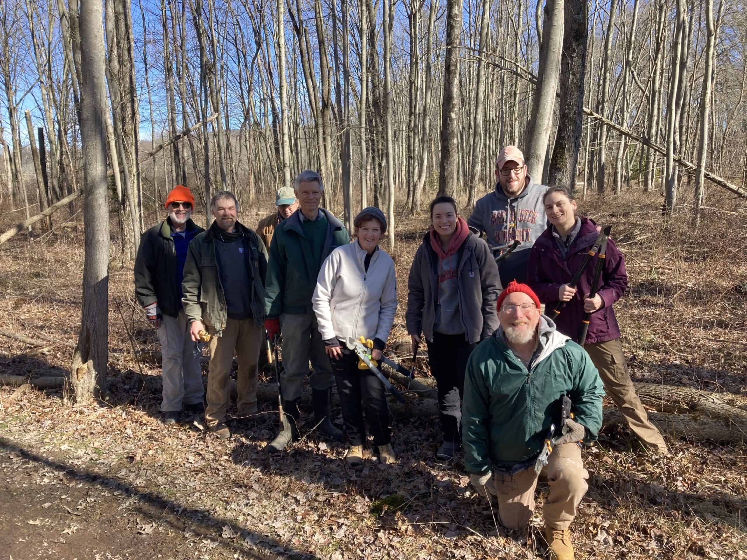 Volunteers with hand tools posing in front of the woods where they were cutting multiflora rose.