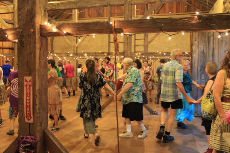 People contra dancing in a barn with twinkling lights above.