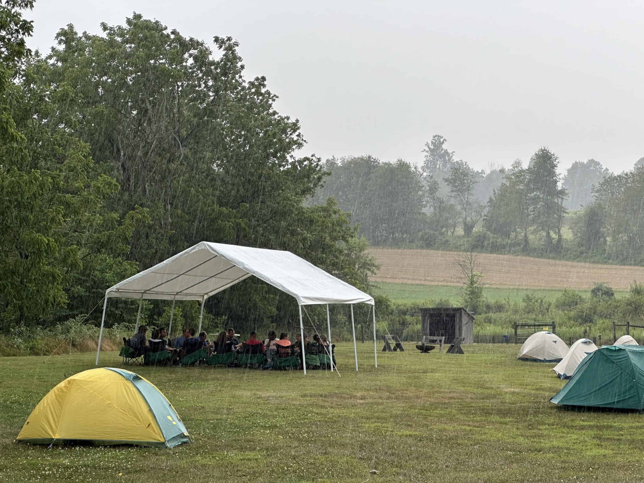 A canopy tent in the rain with people relaxing in chairs underneath it.