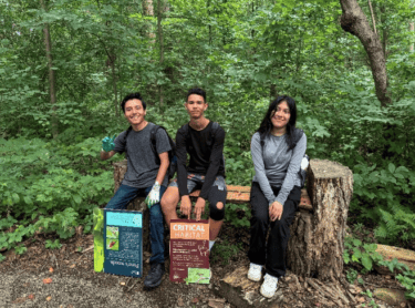 Three youth sit on a bench in the woods with interpretive signs. 