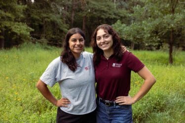 Two young women with Natural Lands staff logos on their shirts stand arm in arm in a meadow.