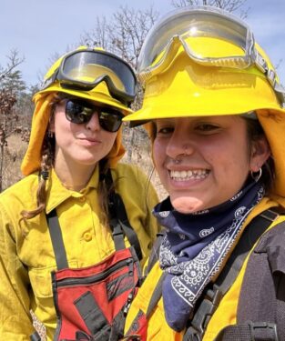 Two young women in yellow protective fire gear smile at the camera